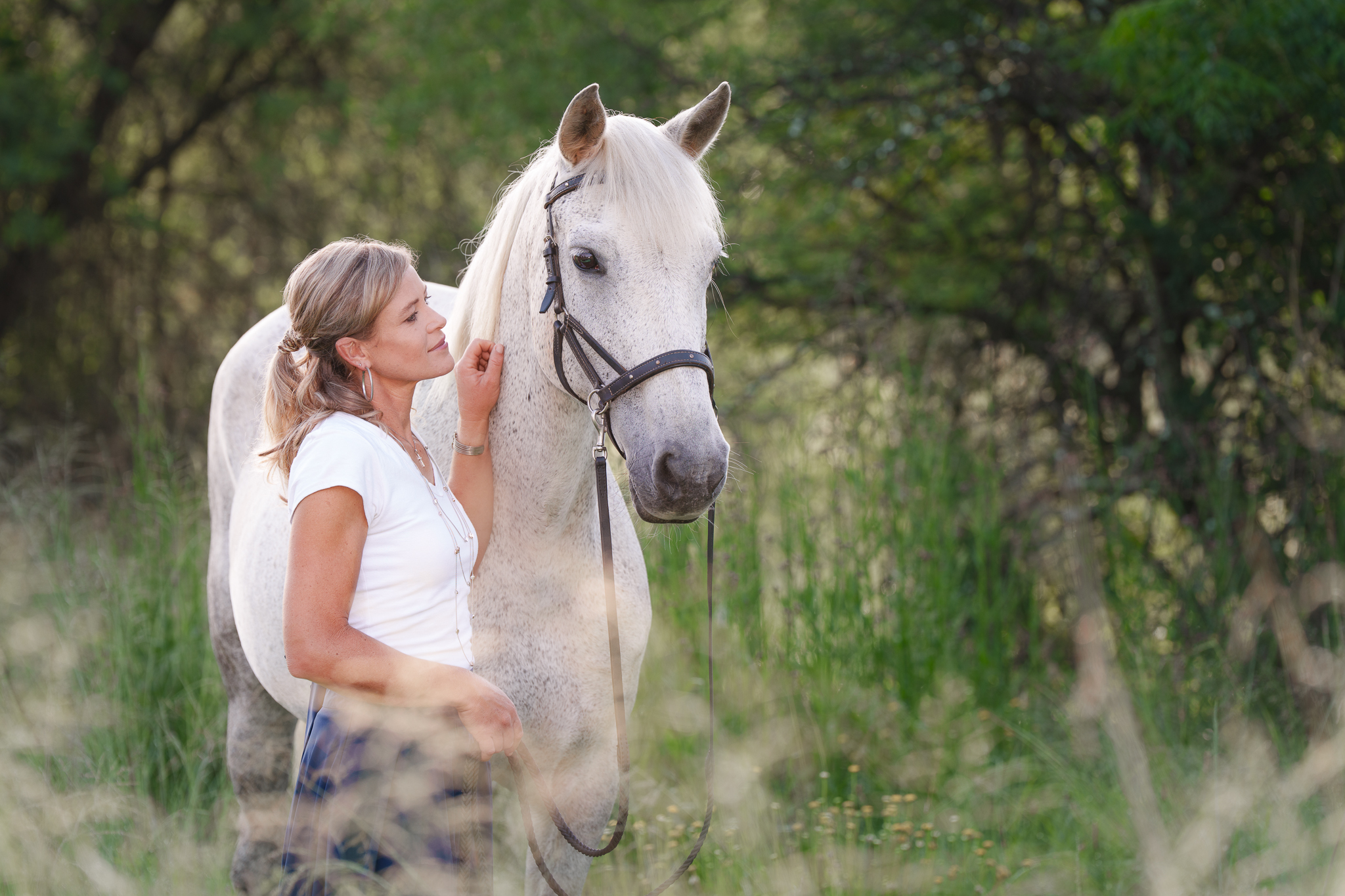 Woman standing beside her white horse in a natural outdoor setting, photographed in soft morning light